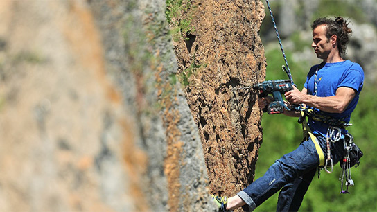 Alpinista w niebieskiej koszulce i dżinsach używa wiertarki akumulatorowej na ścianie skalnej, aby wiercić otwory.