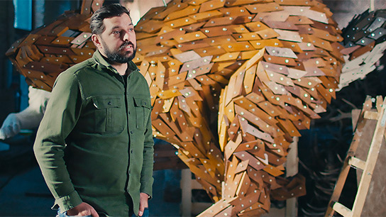 Mężczyzna w oliwkowej koszuli stoi obok dużej, rzeźbiarskiej figury z drewna z wielu kawałków. Figura ma organiczny kształt i utrzymana jest w ciepłych odcieniach drewna. W tle widać inne prace drewniane i warsztatowe otoczenie. Mężczyzna wygląda na zamyślonego i patrzy na rzeźbę.