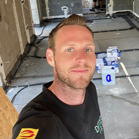 Ein Mann mit kurzem Haar steht in einem Raum mit Werkzeugen und Materialien.