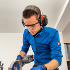 Ein Mann in blauer Kleidung benutzt eine Maschine mit Schutzbrille und Ohrschutz.