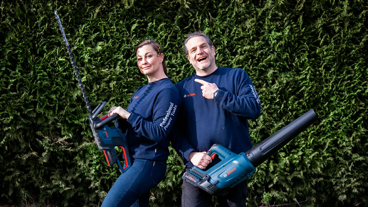 Zwei Personen zeigen Bosch Professional Gartengeräte vor einer Hecke.