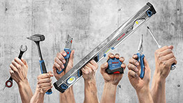 Des mains tiennent divers outils Bosch devant un mur en béton.