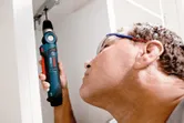 A person wearing safety equipment uses a cordless angle drill to work inside a cabinet.