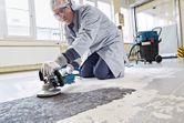 A person wearing safety equipment grinds a concrete floor with a power tool.