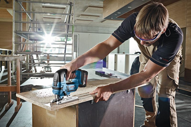 A person wearing safety equipment uses a cordless jigsaw to cut a wooden board.