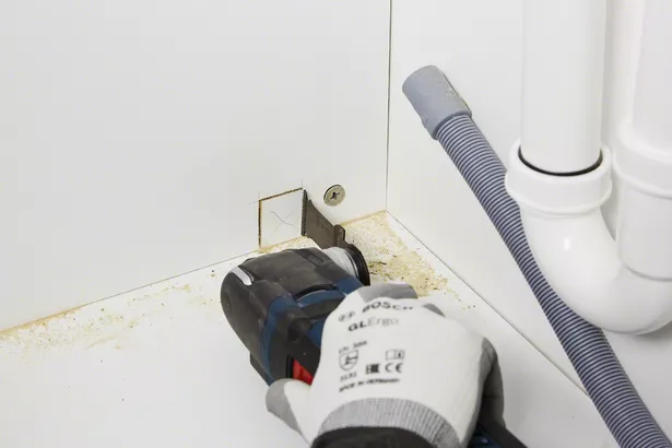 Worker wearing safety equipment cuts a square opening in a cabinet with a power tool.