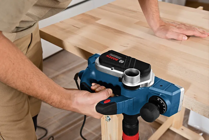 A person uses an electric planer to smooth the edge of a wooden tabletop.