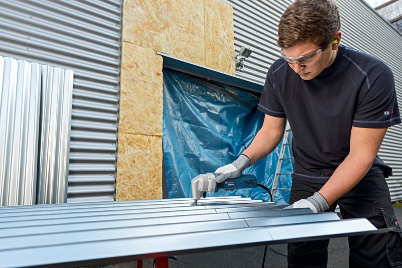 A person wearing safety equipment cuts sheet metal with a nibbler outdoors.