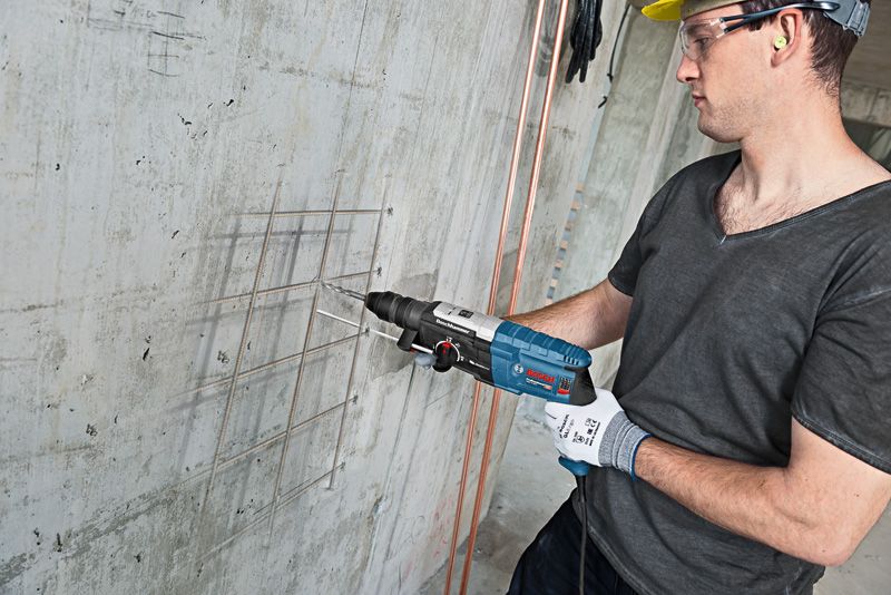 A person wearing safety equipment drills into a concrete wall with a rotary hammer.