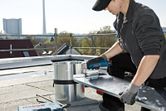 Person wearing safety equipment uses cordless metal shear to cut a sheet on a rooftop.