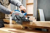 A person guides a circular saw through thick wooden boards in a workshop.