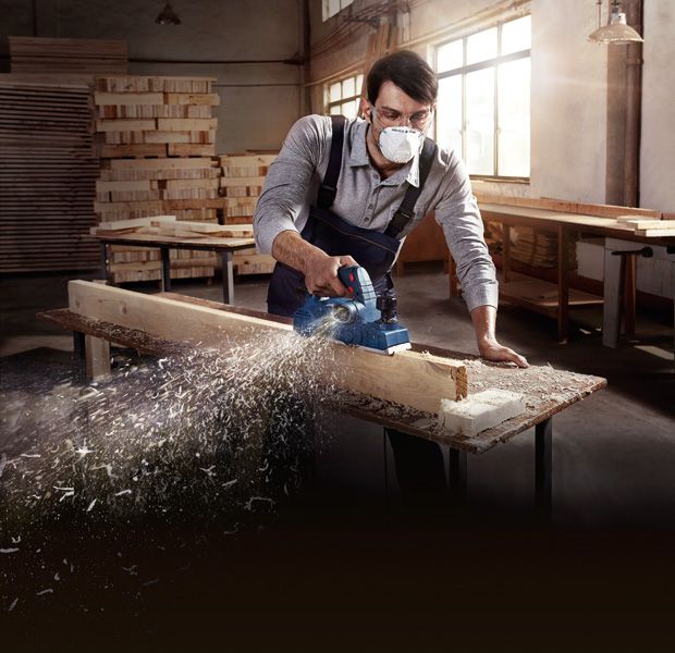 A person wearing safety equipment smooths a wooden plank with a planer.