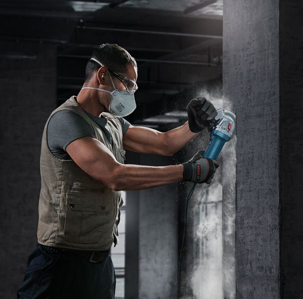A person wearing safety equipment grinds a concrete wall with an angle grinder.