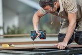 Person wearing safety equipment uses a cordless impact driver to fasten a screw into wood.