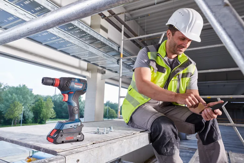 A person wearing safety equipment uses a smartphone beside a cordless drill on a construction site.