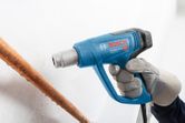 A person wearing safety equipment uses a blue heat gun near a copper pipe.