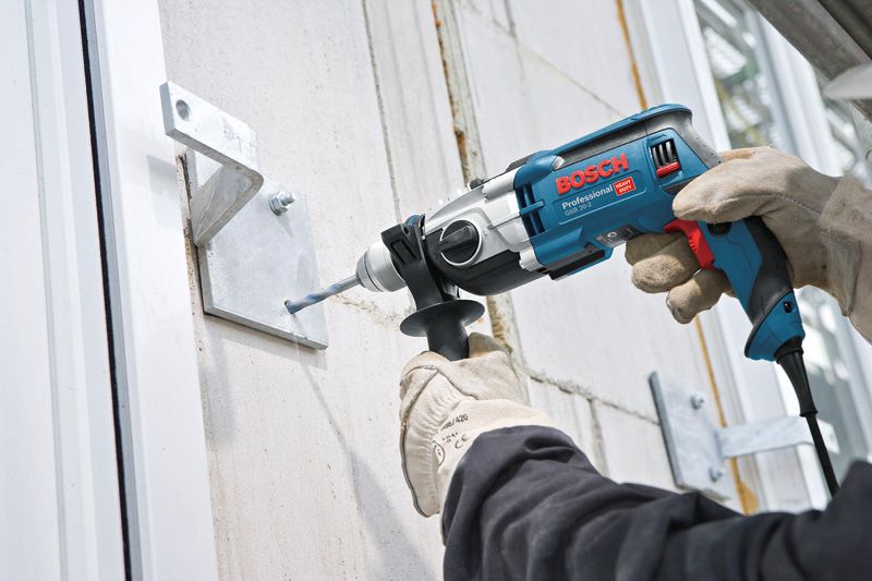 A person wearing safety equipment drills into a metal bracket mounted on a wall.