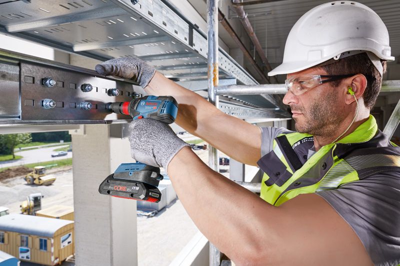 A person wearing safety equipment uses a cordless drill on a steel beam.