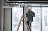 A person wearing safety equipment uses a cordless heat gun on a ladder near ceiling ducts.