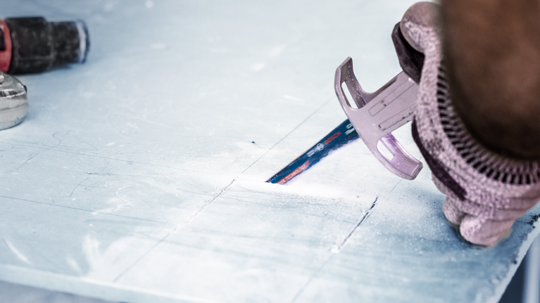 Person wearing safety equipment uses a saw to cut lines in a sheet of material.