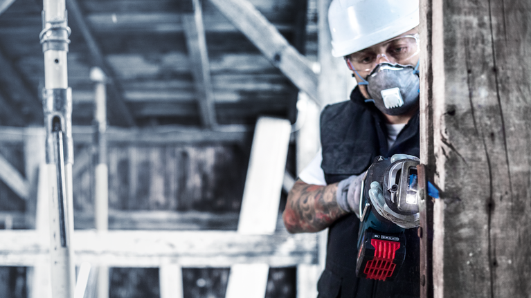 A person wearing safety equipment uses a cordless angle grinder to cut through wood.