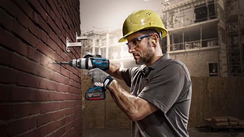 A person wearing safety equipment drills into a brick wall with a cordless power tool.