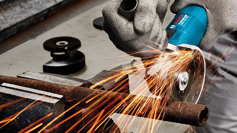 A person wearing safety equipment cuts a metal pipe with an angle grinder, sparks flying.
