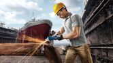 A person wearing safety equipment grinds a metal pipe, producing sparks at a shipyard.
