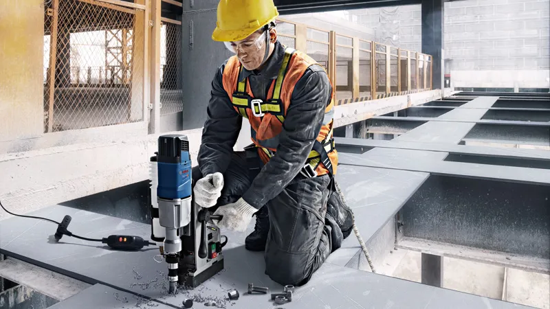 A worker wearing safety equipment operates a magnetic core drill on a steel beam.