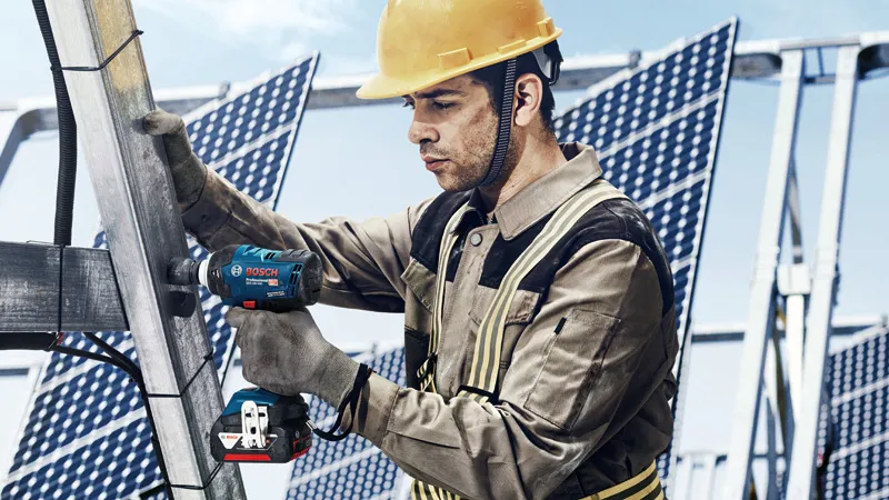 A person wearing safety equipment uses a cordless impact wrench on a metal solar panel frame.