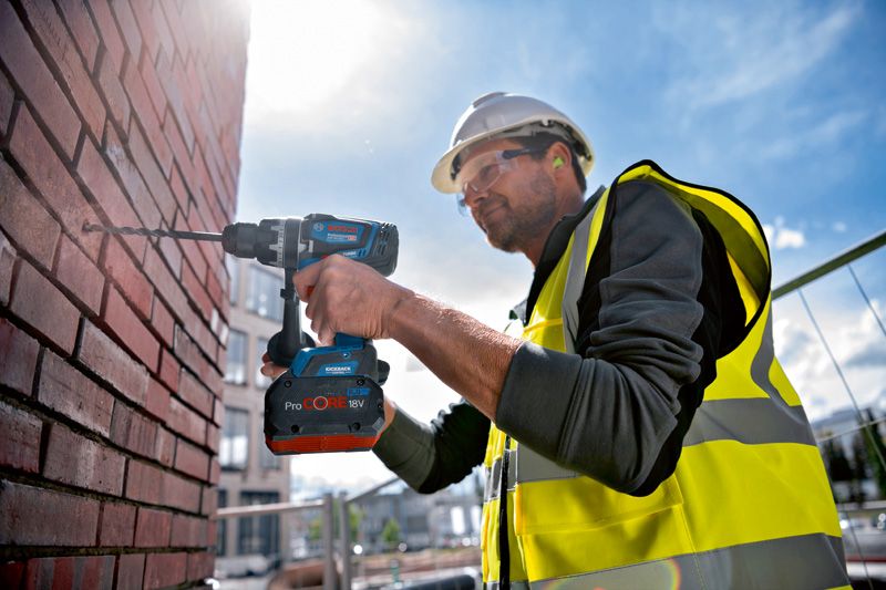 A person wearing safety equipment drills into a brick wall with a cordless combi tool.