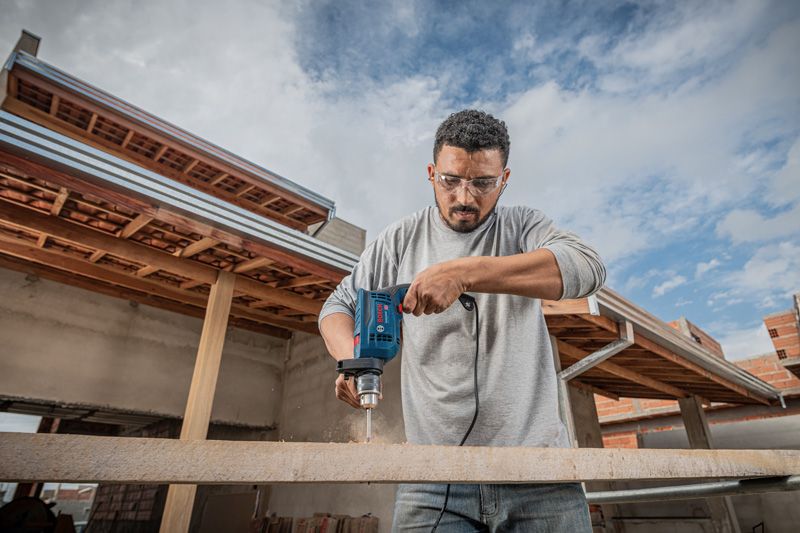 A person wearing safety equipment drills into a wooden beam at a construction site.