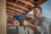 A person wearing safety equipment drills into wood under a wooden roof.
