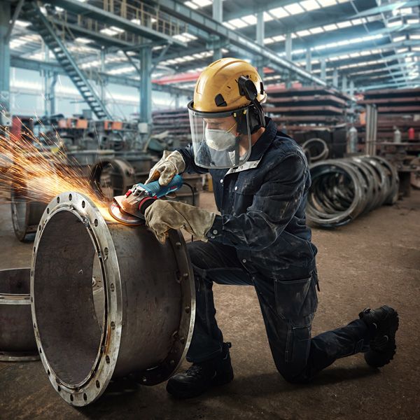 A person wearing safety equipment grinds the edge of a large metal pipe, producing sparks.