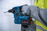 A person wearing safety equipment drills into a tiled wall with a cordless rotary hammer.