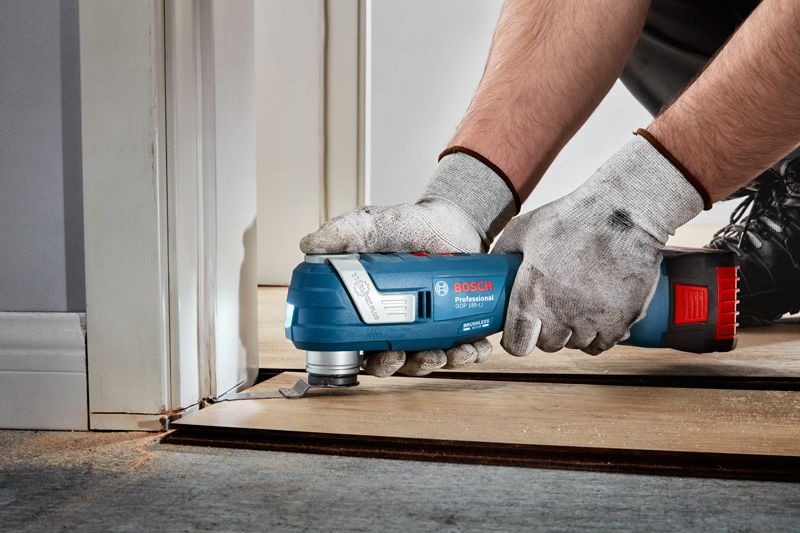 A person wearing safety equipment uses a cordless multi-cutter to trim flooring near a door.