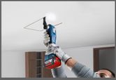 A person wearing safety equipment cuts a ceiling panel with a cordless multi-cutter.