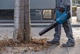 A person wearing safety equipment uses a cordless leaf blower to move leaves near a tree.