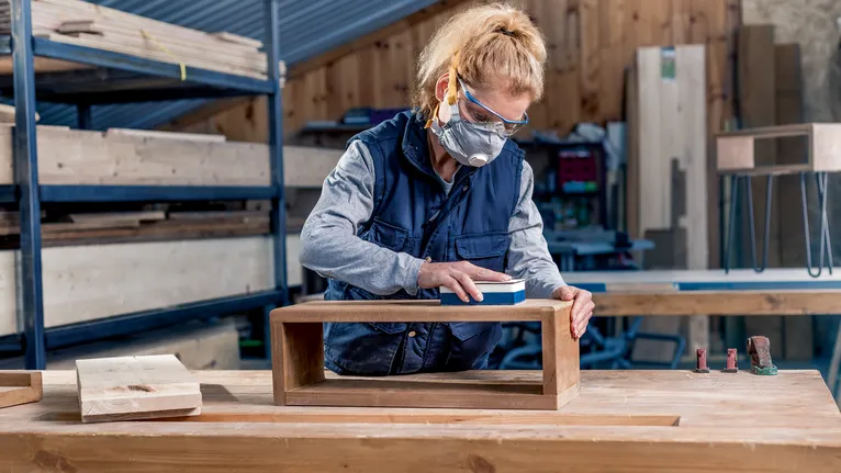 Person wearing safety equipment sands a wooden box in a workshop.