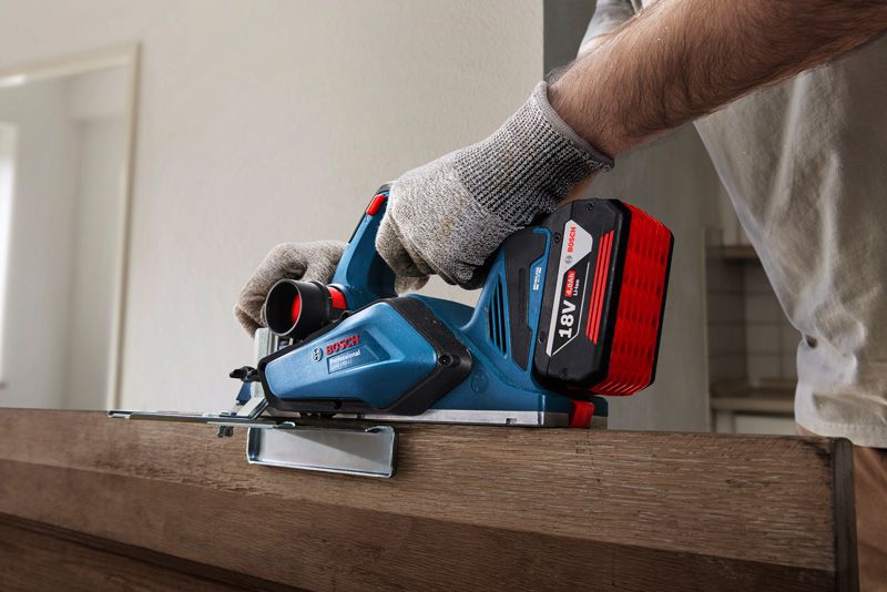 A person wearing safety equipment uses a cordless planer on a wooden door.
