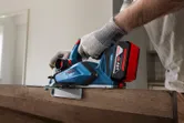 A person wearing safety equipment uses a cordless planer on a wooden door.