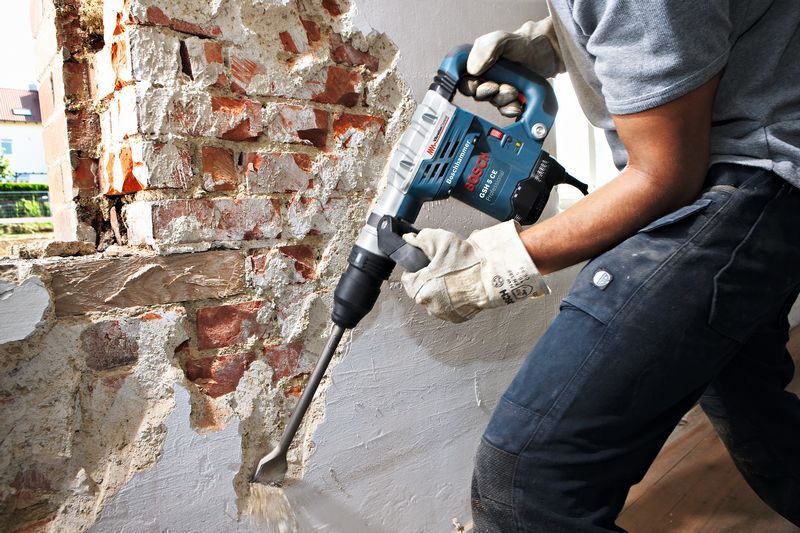 A person wearing safety equipment uses a rotary hammer to remove plaster from a brick wall.