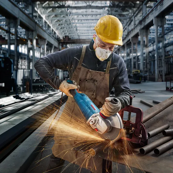 A person wearing safety equipment cuts a metal pipe with a large angle grinder.