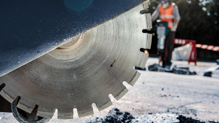 Person wearing safety equipment operates a road saw to cut asphalt pavement.