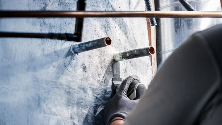 Person wearing safety equipment uses a power tool to cut into a wall around metal pipes.