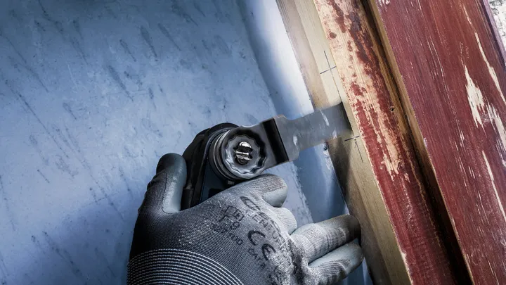 Person wearing safety equipment uses a multi-tool to cut wood near a painted surface.