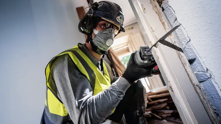 Person wearing safety equipment cuts a doorframe with a reciprocating saw.
