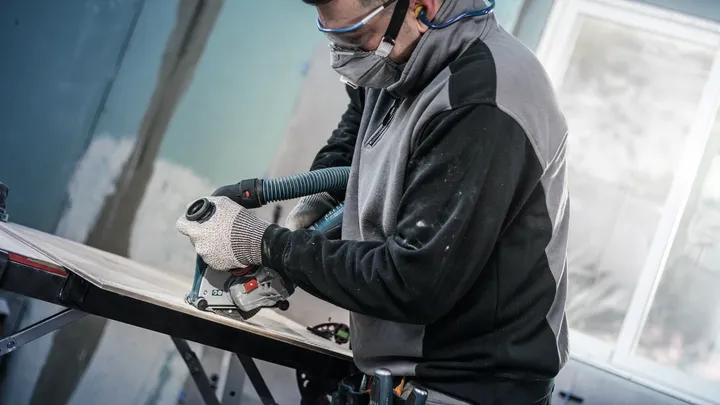 Person wearing safety equipment trims wood with a power tool attached to a vacuum hose.