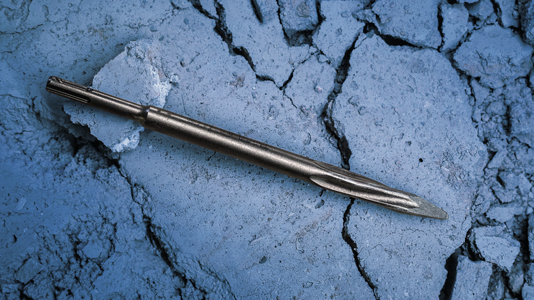Metal chisel bit lying on cracked concrete surface.