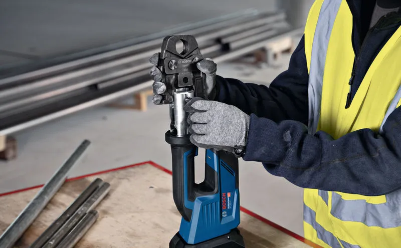 A person wearing safety equipment uses a cordless press tool at a workbench.